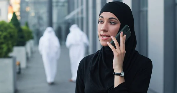 Портретний вигляд серйозної арабської бізнес-леді в шарфі голови, що стоїть на вулиці і спілкується через смартфон зі своїми діловими партнерами — стокове фото