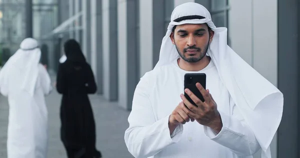 Utilizzando smartphone. Vista ritratto in vita dell'uomo d'affari arabo che scrive un messaggio serio a qualcuno mentre tiene il cellulare per strada — Foto Stock