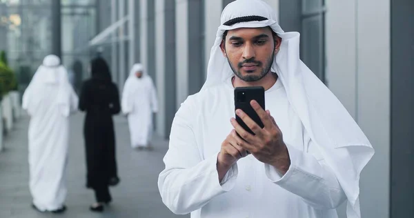 Usando smartphone. Vista de la cintura hacia arriba del hombre de negocios árabe escribiendo un mensaje serio a alguien mientras sostiene su teléfono celular en la calle —  Fotos de Stock