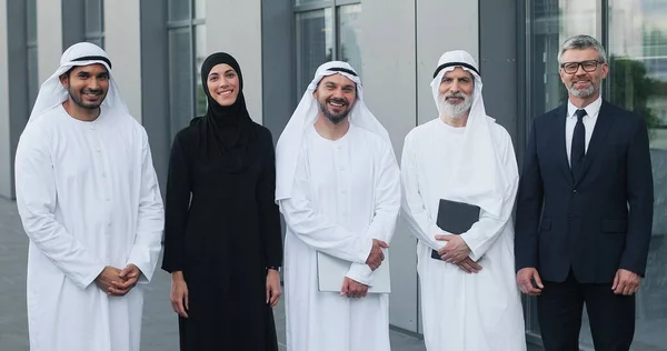 Un equipo exitoso. Cintura hacia arriba retrato vista de la gente de negocios árabe de pie con su socio de negocios caucásico y sonriendo a la cámara mientras posan al aire libre —  Fotos de Stock