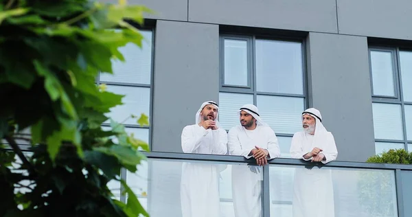 Volledig uitzicht op de drie saudi sjeik in kanduras gebaren en bespreken van iets terwijl ontspannen op het balkon tijdens hun pauze. Bedrijfsconcept — Stockfoto