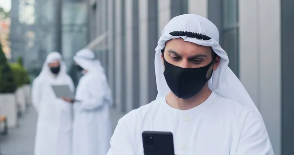 Використання смартфона. Прочекай портрет арабського бізнесмена, який носить захисну маску, пишучи серйозне повідомлення комусь, тримаючи свій мобільний телефон на вулиці. Пандемічна концепція — стокове фото