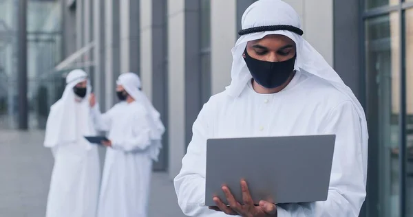 Vista verticale in vita dell'uomo arabo che indossa una maschera protettiva che lavora da solo con il suo computer portatile durante la pandemia. Business e covid 19 concetto — Foto Stock