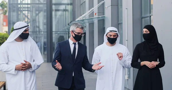 Na schůzce. Plný pohled na čtyři muslimské kolegy v ochranných maskách procházející ulicí v blízkosti obchodního centra a chatování — Stock fotografie