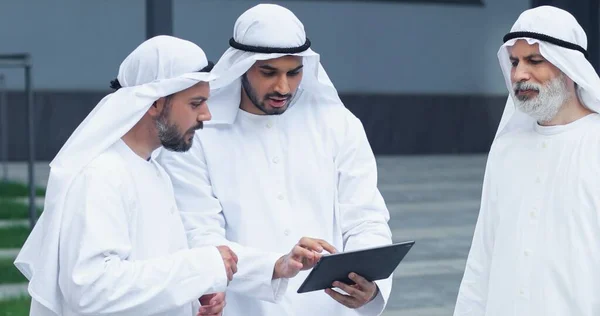 Vista del retrato de la cintura hacia arriba del equipo de empresarios árabes charlando entre sí y mirando algo en la tableta mientras están de pie en la calle. Concepto empresarial —  Fotos de Stock
