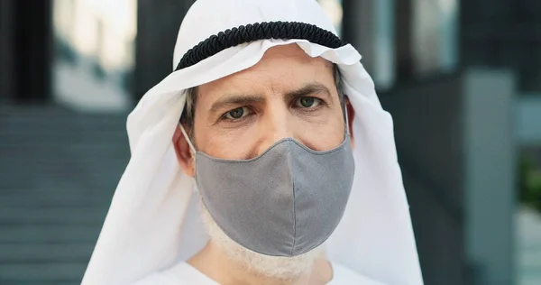 Waist up portrait view of the serious senior businessman wearing kandura and protective mask standing at the street at the break and looking at the camera. Coronavirus pandemic concept — Stock Photo, Image