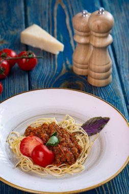 Beyaz tabakta spagetti bolonez makarna, tuz, biber, peynir ve domates mavi ahşap arka planda.