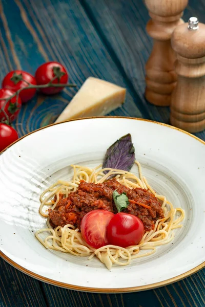 Beyaz tabakta spagetti bolonez makarna, tuz, biber, peynir ve domates mavi ahşap arka planda.