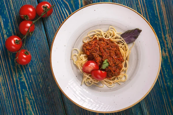 Beyaz tabakta spagetti bolonez makarna, tuz, biber, peynir ve domatesler mavi ahşap arka planda.