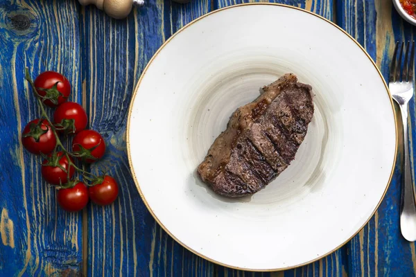 Kızarmış biftek, kiraz domatesleri, mavi ahşap masada tuz ve biber..