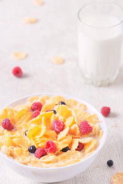 Bir kase mısır gevreği, sütlü, taze ahududu, yabanmersini. Sağlıklı kahvaltı için bir sürahi süt ve beyaz tahta üzerinde bir bardak.