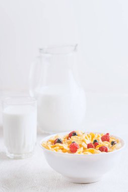 Bir kase mısır gevreği, sütlü, taze ahududu, yabanmersini. Sağlıklı kahvaltı için bir sürahi süt ve beyaz tahta üzerinde bir bardak.