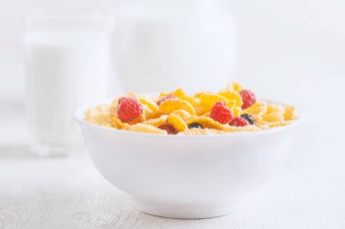 Bir kase mısır gevreği, sütlü, taze ahududu, yabanmersini. Sağlıklı kahvaltı için bir sürahi süt ve beyaz tahta üzerinde bir bardak.