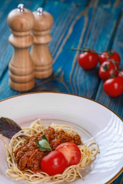 Beyaz tabakta spagetti bolonez makarna, tuz, biber, peynir ve domates mavi ahşap arka planda.