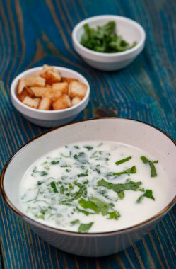 Yoğurt çorbası, mavi ahşap bir masada otlar ve krakerler.