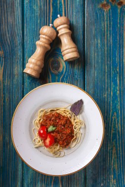 Beyaz tabakta spagetti bolonez makarna, tuz, biber, peynir ve domatesler mavi ahşap arka planda.