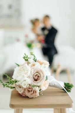 Bir buket bej ve beyaz gül, ön planda şakayıklar ve arka planda bulanık bir düğün çifti.