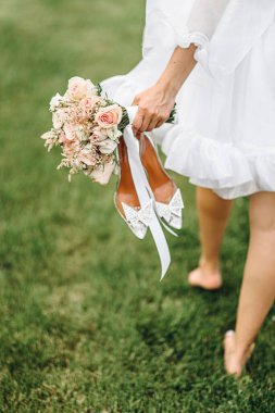 Çıplak ayaklı bir kız, elinde ayakkabılar ve bir buket çiçekle çimenlerde yürüyor.