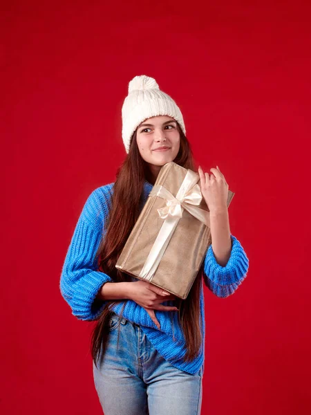 Dívka v modrém svetru a bílý klobouk drží dárek v rukou na červeném pozadí Royalty Free Stock Obrázky