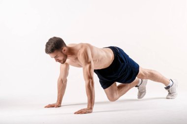 Spor fiziğinde sakallı genç bir adam beyaz bir arka planda yerden şınav çekiyor.