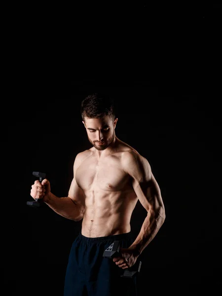 Um homem com um fundo negro com halteres nas mãos. jovem atleta barbudo. — Fotografia de Stock