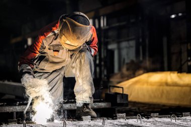 A young  man welder in  uniform, welding mask and welders leathers, weld  metal  with a  welding machine at the construction site, yellow sparks fly to the sides clipart