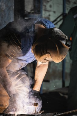 Güçlü bir adama yaklaşan üniformalı bir kaynakçı, kaynak maskesi ve kaynakçı derisi, bir metal ürün inşaat alanında kaynak makinesiyle kaplıdır.