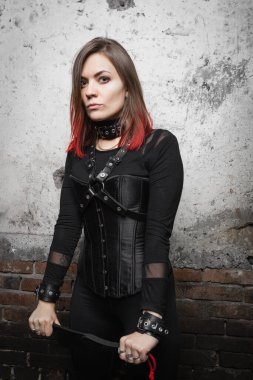 A  attractive dominant woman with piercings and bright hair in a black corset, with leather harnesses and bracelets, holds a belt in her hands, looks sternly against the background of an old wall. clipart