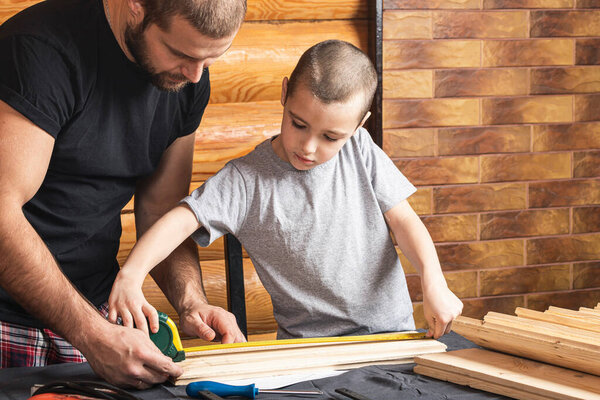 Father and son together make a wooden birdhouse in the workshop. Cheerful father with little boy measure with tape measure wooden planks on table in workshop