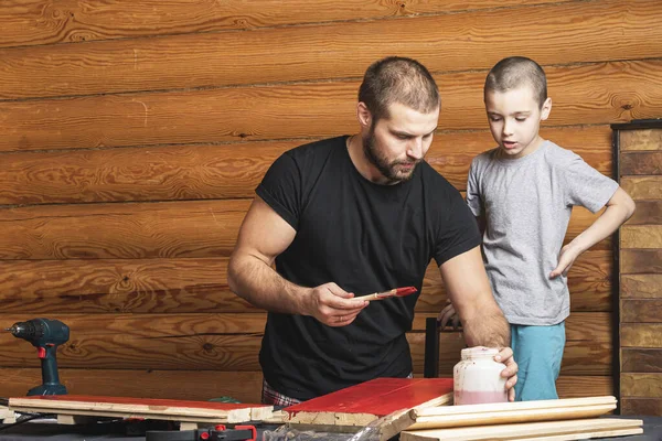 Father and son together make a wooden birdhouse in the workshop. Cheerful father with a little boy paint a wooden board with a brush in red  on a table in the workshop
