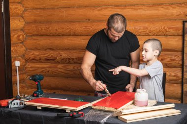 Babası elinde kırmızı boyayla bir fırça tutuyor ve tahta bir yüzey boyuyor, oğluna resim yapmayı öğretiyor, açık bir boya kutusu ve fırçanın yanında. DIY kavramı.