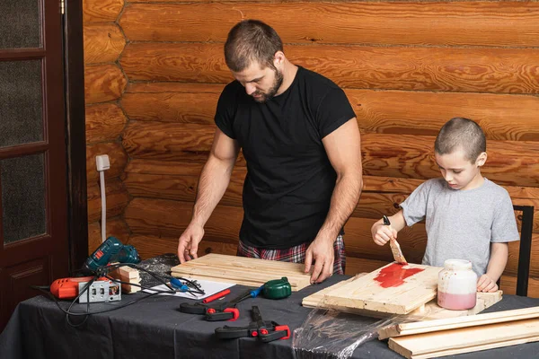 Dad and son are   painting a wooden board with a brush in red,  how to build a bird house, tools and a beam on the table in the workshop. Carpentry training concept for kids