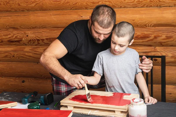 Dad holds a brush with red paint in his hand and paints a wooden surface, teaches his son to paint, next to an open can of paint and a brush. DIY concept.