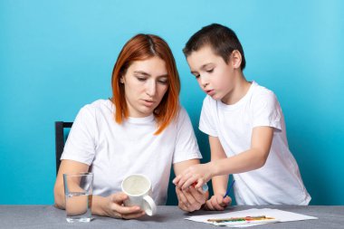 Beyaz tişörtlü anne ve oğlu boyayla meşgul, mavi arka planda bir kupa kırıyorlar. Anaokulunda çizim yapmanın temellerini öğrenmek.