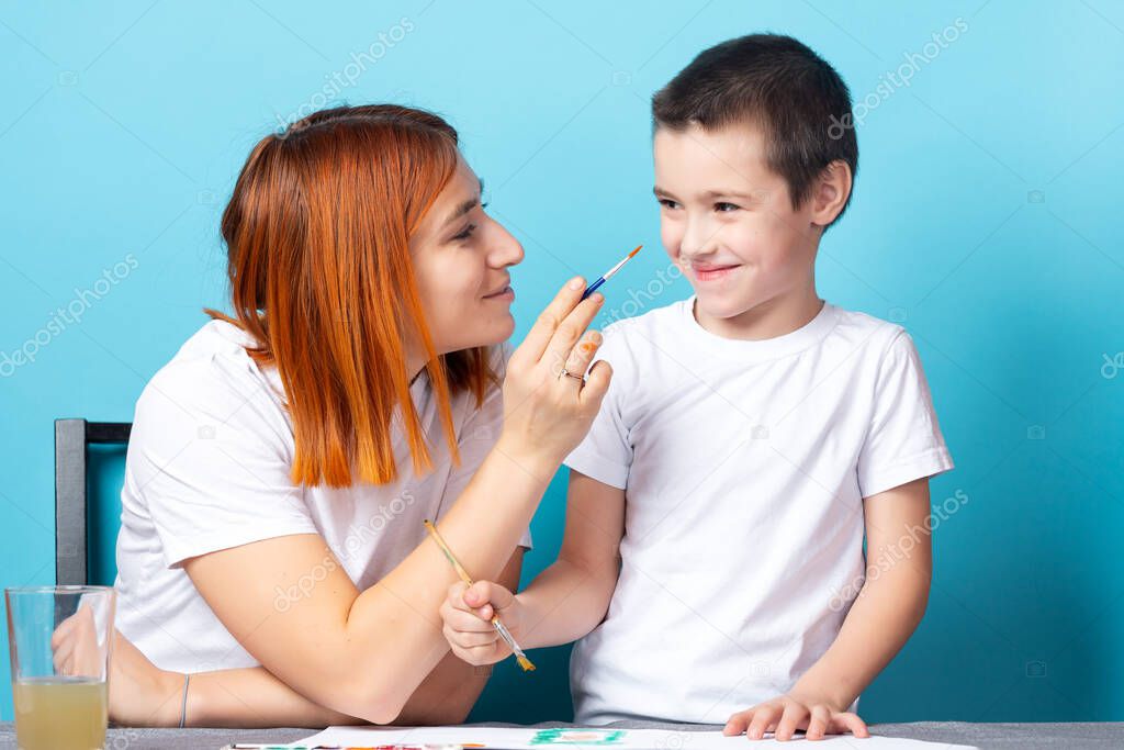 Children's creativity. Mother and son paint and having fun, mom paints a nose for her son with a brush on blue background