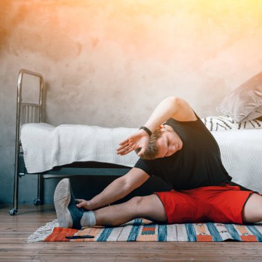 Genç bir adam spor için eve gidiyor, online antrenman. Sporcu esniyor, meditasyon yapıyor, yatak odasında bir sicimin üzerinde oturuyor, arka planda bir yatak, bir halı var..