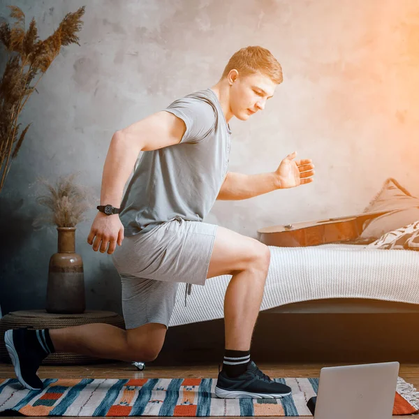 若い男が家でスポーツのために行く ノートパソコンからオンラインワークアウト 背景にはベッド カーペットがあります 彼らはベッドの上に座っています — ストック写真