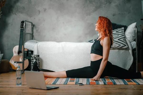 The beauty woman goes in for sports at home. Cheerful sporty woman with red hair stretches to the leg  and watches  in laptop, shootting  blog  in the bedroom