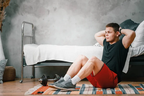 Young man goes in for sports at home, training online. The athlete makes the press, smiles,  on background bed, vase, carpet .