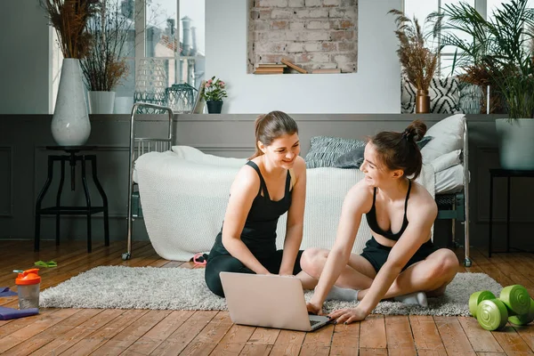 Young women go in for sports at home, workout online. Two athletes are stretching, meditating, discussing workout in the bedroom, in the background there is a bed, a vase, a carpet.