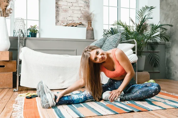A young woman goes in for sports at home, online workout . The athlete  stretching , meditating, sitting on a floow in the bedroom, in the background there is a bed, a vase, a carpet.