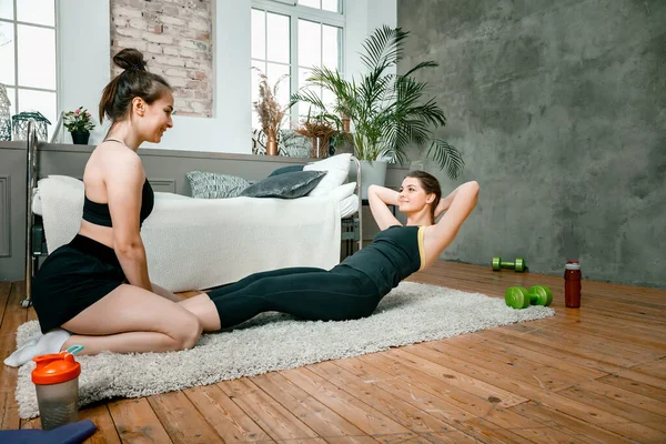 Two women friends at home chatting, smiling and makes sports. Cheerful athletic woman presses, and the second sits nearby and monitors correct execution in the bedroom