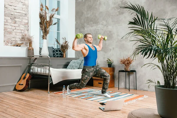 A young man goes in for sports at home, online workout from the laptop. The athlete makes lunges with dumbells  in the bedroom, in the background there is a bed, a vase, a carpet.