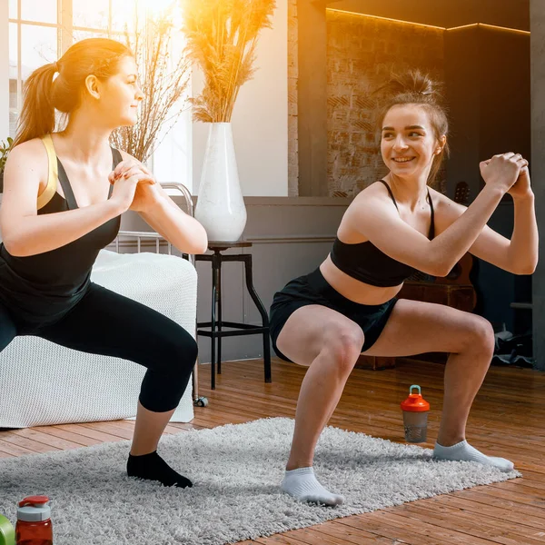 Young women go in for sports at home, workout online. Two athletes are makes squat   in the bedroom, in the background there is a bed, a vase, a carpet.