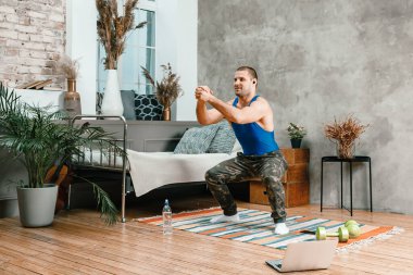 Genç bir adam evde spor yapıyor, bilgisayardan antreman yapıyor. Sporcu çömelir, yatak odasında film izler, arka planda bir yatak, bir vazo, bir halı vardır..