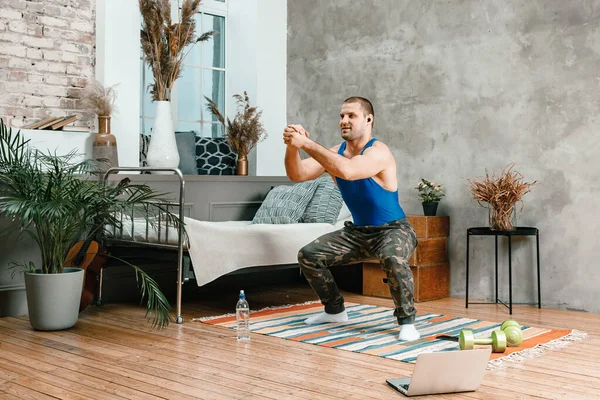 A young man goes in for sports at home, online workout from the laptop. The athlete makes squat , watches a movie  in the bedroom, in the background there is a bed, a vase, a carpet.