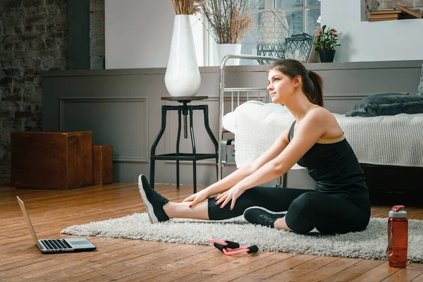 A young woman goes in for sports at home, online workout . The athlete  stretching , meditating, sitting on a floor  in the bedroom