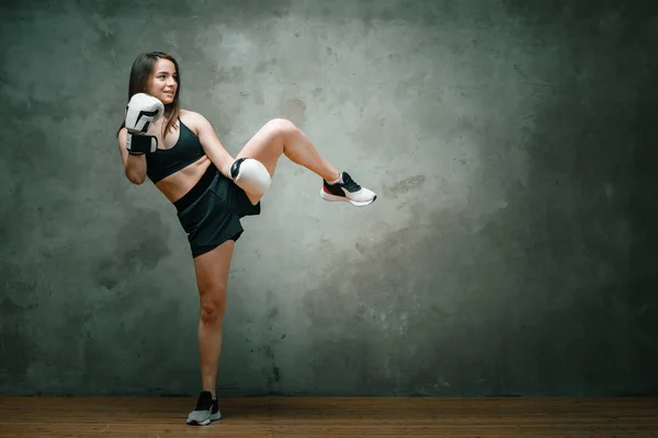 Young athletic woman boxer in shorts, short top and boxing gloves doing a kick above his head on dark gray background. Full length. Wall in background.