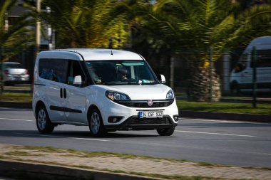 Alanya, Türkiye 12 Nisan 2021: Beyaz Fiat Doblo sıcak bir yaz gününde palmiyelerin arkasına doğru hızla sürüyor 