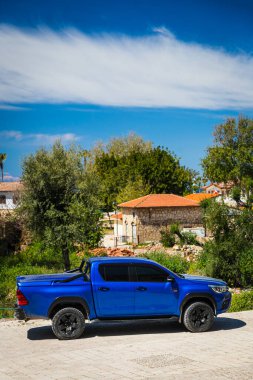Yan, Türkiye 15 Nisan 2021: Mavi Toyota Hilux sıcak bir yaz gününde eski sokak, ağaçlar, palmiyeler karşısında sokağa park etti 